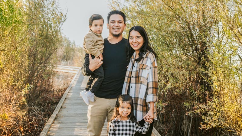 Oregon State University student Esteban Contreras stands with his wife and two young children on a wooden walkway surrounded by tall, green foliage.